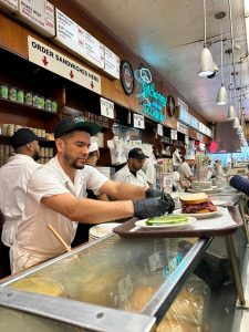 Ein Mitarbeiter von Katz´s Delicatessen bereitet das Essen an der Theke vor. Man sieht das beliebte Pastrami-Sandwich auf dem Tablett stehen.